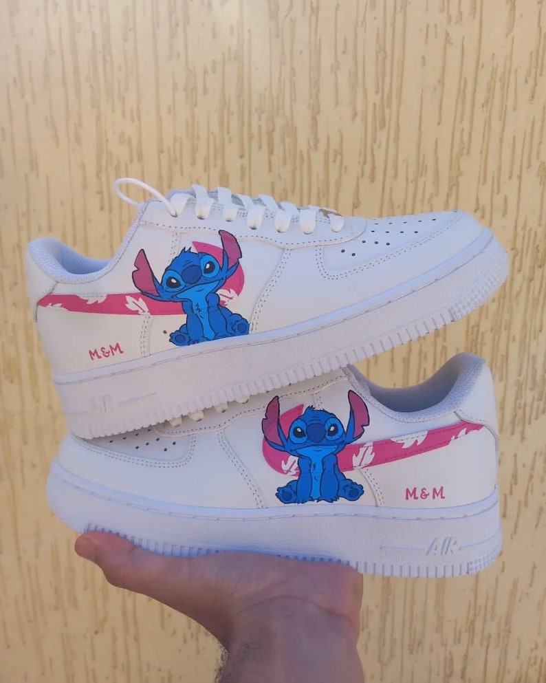 May include: White sneakers featuring a blue cartoon character with large ears and a pink accent. The text "M&M" is printed in pink on the side of the shoe. The shoes have white soles and laces.