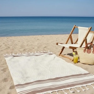 May include: A beach scene with a white and brown patterned beach towel laid out on the sand. A wooden beach chair, straw tote bag, and a drink with a lime wedge are nearby. The ocean is in the background.