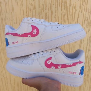 May include: White sneakers with pink leaf-patterned swooshes and the text "05/05". The design includes a blue cartoon character on the side. The shoes have white soles and laces.