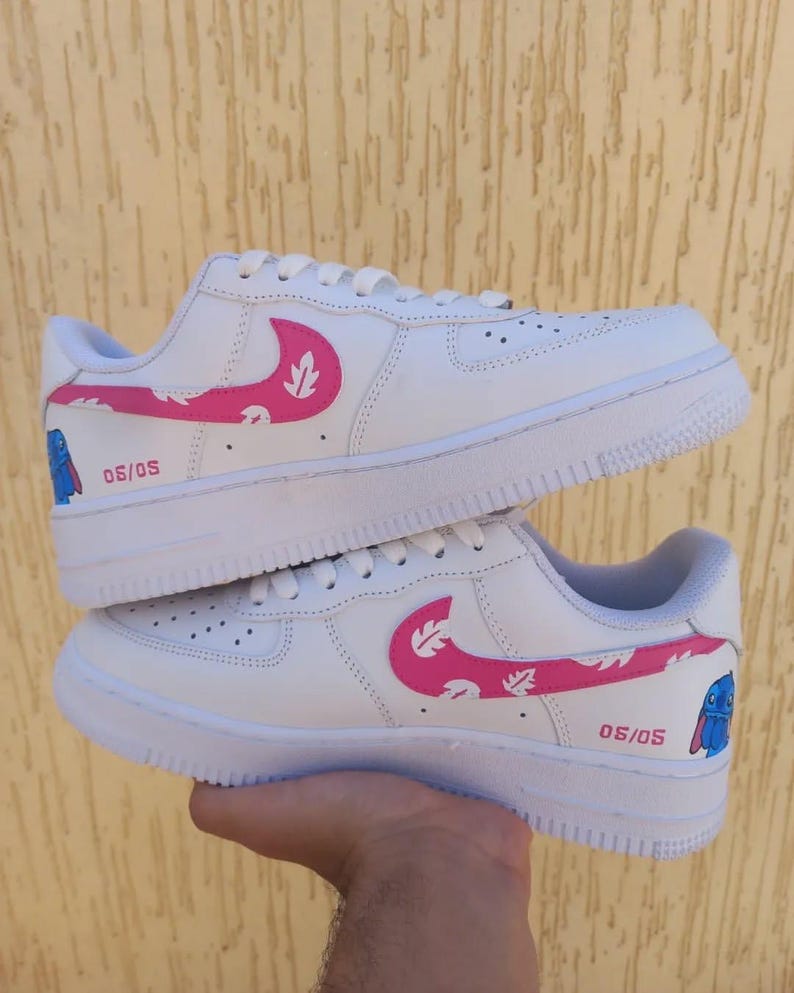 May include: White sneakers with pink leaf-patterned swooshes and the text "05/05". The design includes a blue cartoon character on the side. The shoes have white soles and laces.