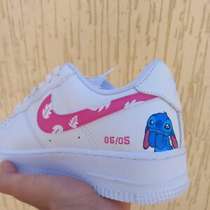 May include: White athletic shoe with a pink swoosh featuring a leaf design. The shoe has a cartoon character illustration and the text "05/05" in pink. The shoe is held by a hand.
