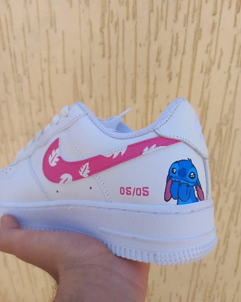 May include: White athletic shoe with a pink swoosh featuring a leaf design. The shoe has a cartoon character illustration and the text "05/05" in pink. The shoe is held by a hand.