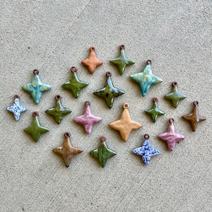May include: A collection of seventeen four-pointed star-shaped ceramic pendants in various colors including olive green, tan, pink, and blue-and-white speckled patterns. Each star features a small circular loop at the top for hanging or attachment.