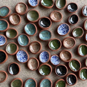 May include: A collection of small, handmade ceramic bowls with a rustic, terracotta-colored rim. The bowls feature a variety of interior colors, including blue, green, brown, and black, some with speckled patterns. The bowls are arranged closely together.