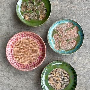 May include: Four circular ceramic grater plates with textured centers. One is pink with a dotted pattern, one is green with a textured abstract shape, one is blue and green with a textured shape, and one is green with a circular textured center.