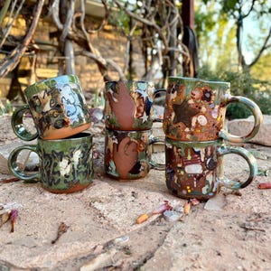 May include: Five ceramic mugs with varied hand-painted designs. Two mugs feature dark green glaze with black abstract patterns. Two mugs display brown hand shapes and white stars on a green background. One mug shows a brown star and colorful dots.