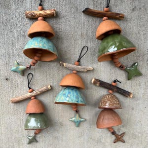May include: Handmade ceramic wind chimes in various colors, including teal, green, and brown. Each chime features a star-shaped accent and is suspended from a wooden branch. The chimes are designed with a rustic, natural aesthetic.