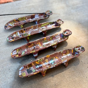 May include: Four elongated, brown ceramic incense holders arranged in a row. Each holder features a glossy finish with colorful, abstract painted patterns in yellow, green, blue, and white. They have small legs and a circular end for incense.