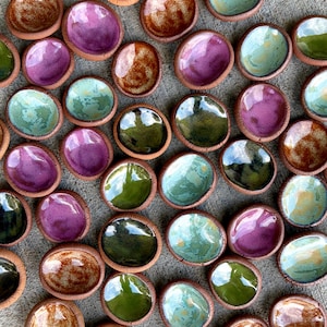 May include: A collection of small, round, hand-formed ceramic dishes with unglazed terracotta edges and glossy, multi-colored glazed interiors in shades of purple, green, blue, and brown, arranged in a scattered pattern on a gray surface.