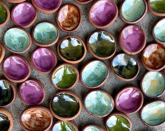 May include: A collection of small, round, hand-formed ceramic dishes with unglazed terracotta edges and glossy, multi-colored glazed interiors in shades of purple, green, blue, and brown, arranged in a scattered pattern on a gray surface.