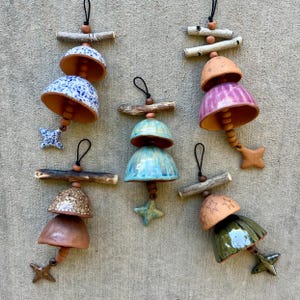 May include: Five hanging ceramic bell ornaments suspended from wooden sticks. Each bell features unique glazes including blue speckled, pink striped, solid brown, turquoise, and green with star patterns, all finished with small star-shaped clappers.