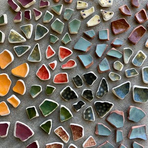 May include: A collection of irregularly shaped ceramic pieces in various colors, including orange, red, green, and blue. The pieces have a textured appearance and are arranged on a gray surface. Some pieces have a speckled or patterned design.