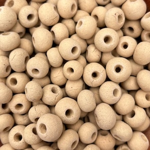 May include: A close-up shot of a pile of beige, round beads with a hole through the center. The beads have a rough, textured surface and vary slightly in size and shape. These beads are suitable for jewelry making and crafting.