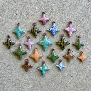 May include: A collection of seventeen four-pointed star-shaped ceramic pendants. Each piece features a small hole at the top for hanging. The pendants display a variety of finishes, including solid matte earth tones, glossy blue, green, and pink glazes.