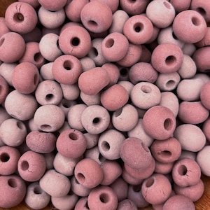 May include: Close-up of a pile of pink and light pink ceramic beads. Each bead is round with a hole through the center. The beads have a matte finish and vary slightly in color, creating a textured appearance. Ideal for jewelry making and crafts.