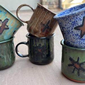 May include: A stack of six ceramic mugs in various colors, including teal, brown, blue, and green. Each mug features a unique hand-painted design, such as a star or abstract shapes. The mugs have a rustic, handcrafted appearance.