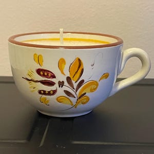 May include: A white teacup candle with a floral design in yellow, brown, and maroon. The rim of the cup is brown, and the candle wax is a light yellow. The teacup has a handle and sits on a dark surface.