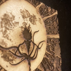 Puede incluir: Una obra de arte pirograbada que presenta una luna llena con una silueta oscura y detallada de una criatura. La luna est&aacute; rodeada por un dise&ntilde;o de sol, todo en tonos marrones y beige.