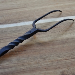 Könnte beinhalten: Eine schwarze, handgeschmiedete Metall-Bratengabel mit gedrehtem Griff und zwei gebogenen Zinken. Die Gabel ist vor einem hölzernen Hintergrund platziert und zeigt ihr rustikales Design und ihre Handwerkskunst.