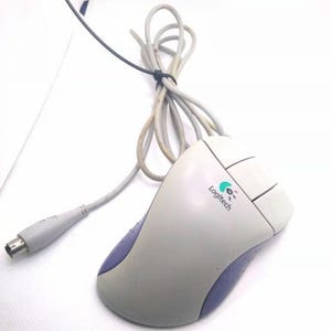 May include: A light gray and purple wired computer mouse featuring the Logitech logo printed on the top surface. The device includes two primary buttons, a scroll wheel, and a long gray cable ending in a round PS/2 connector for computer input.