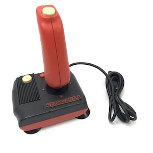 Może przedstawiać: Czerwono-czarny joystick Starmaster z wysokim czerwonym uchwytem z kremowym przyciskiem na górze, czarną podstawą z kremowym przyciskiem bocznym, czerwonymi strzałkami kierunkowymi oraz czarnym zwijanym kablem z wtyczką połączeniową.