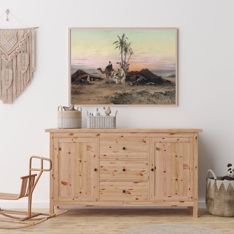May include: A light wood dresser with three drawers and two doors. The dresser has black knobs on the drawers and doors. A painting of a desert scene with camels and people is hanging on the wall above the dresser.