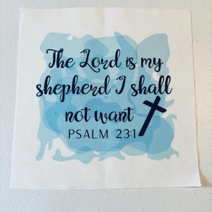 May include: A square white fabric patch featuring a light blue watercolor-style background. The text reads, The Lord is my shepherd I shall not want PSALM 23:1, printed in a dark navy blue script font, accompanied by a small navy blue cross icon.