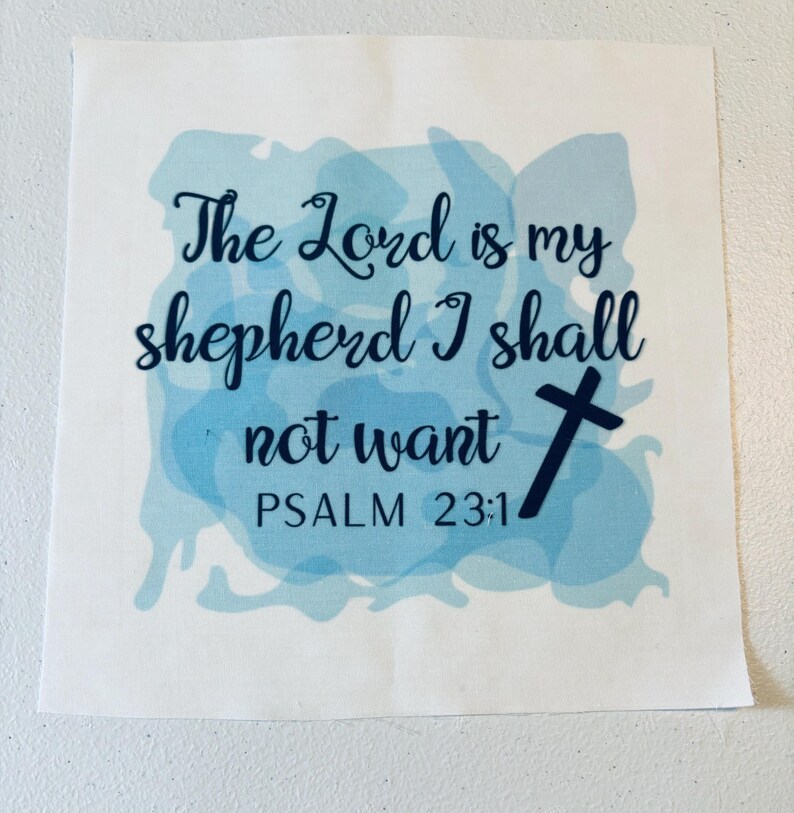 May include: A square white fabric patch featuring a light blue watercolor-style background. The text reads, The Lord is my shepherd I shall not want PSALM 23:1, printed in a dark navy blue script font, accompanied by a small navy blue cross icon.
