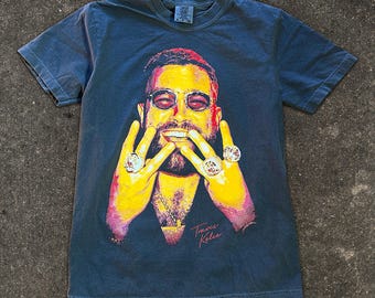 May include: A charcoal gray short-sleeve t-shirt featuring a vibrant, multi-colored graphic print of a bearded individual wearing round sunglasses and displaying large rings on fingers. The name Travis Kelce is printed in a cursive pink font.
