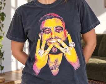 以下が含まれることがあります： チャコールグレーの半袖Tシャツ。サングラスをかけ、指に大きな指輪をいくつもはめた髭のある人物の鮮やかなマルチカラーのグラフィックプリントが施されており、その人物は顔の近くで手を掲げるポーズをとっている。