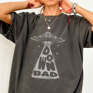 May include: An oversized t-shirt in a dark grey colour with a graphic of a UFO and the text "DOWN BAD" printed on the front.