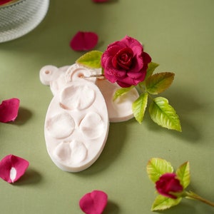May include: A pink silicone mold for making polymer clay rose petals, with a single red rose and green leaves on a green background.