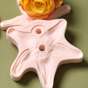 Puede incluir: Un molde de silicona rosa en forma de estrella de cinco puntas con dos agujeros en el centro. El molde está etiquetado como "Zara Sweet Art".
