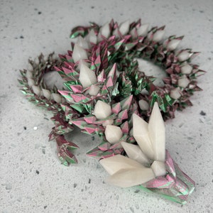 Puede incluir: Una escultura de dragón de origami impresa en 3D con un acabado metálico rosa y verde. El dragón está enroscado con la cabeza apoyada en el suelo. La cabeza del dragón tiene púas blancas de aspecto cristalino.