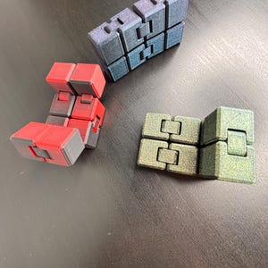 Könnte beinhalten: Drei Fidget Toys in verschiedenen Farben: Rot, Blau und Grün. Jedes Spielzeug ist aus Kunststoff und hat eine strukturierte Oberfläche. Die Spielzeuge sind in Dreiecksform auf einer dunklen Oberfläche angeordnet.