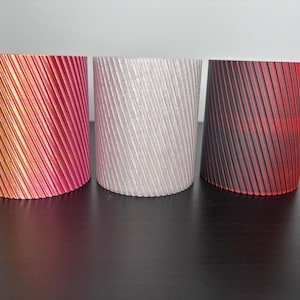 Könnte beinhalten: Drei zylindrische dekorative Behälter mit einem geriffelten, diagonalen Muster. Die Behälter sind in den Farbtönen Rosa, Silber und Schwarz mit roten Akzenten gehalten. Die Objekte werden auf einer dunklen Oberfläche präsentiert.