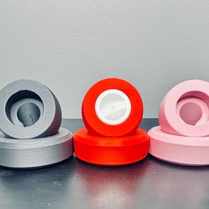 May include: Five different colored 3D printed round objects with a hole in the center. The objects are white, gray, red, pink, and black.