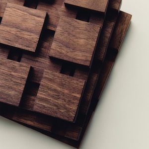 May include: A stack of square dark brown wooden coasters featuring a grid pattern of raised blocks and recessed channels. The natural wood grain is visible on the surface of each square piece, creating a textured, geometric appearance.