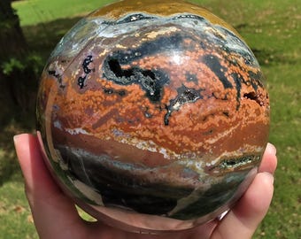 May include: A polished spherical stone featuring a complex pattern of earthy colors including deep brown, rust orange, black, and cream. The surface displays natural mineral inclusions, swirling bands, and small crystalline cavities throughout.