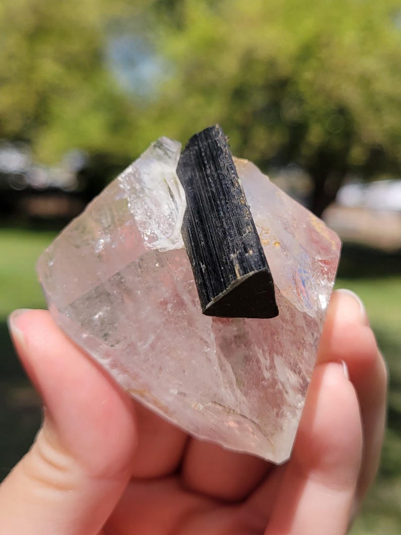 May include: A clear quartz crystal with a black tourmaline crystal embedded in it. The quartz is a pale pink color and has a faceted shape. The black tourmaline is a dark, opaque color and has a rough texture. The crystals are held in a hand.