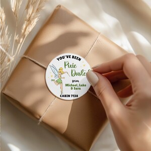 May include: A round white sticker featuring a cartoon fairy with blonde hair and green clothing, surrounded by yellow sparkles. The text reads: YOU'VE BEEN Pixie Dusted from Michael, Luke & Sara CABIN 7138. The sticker is applied to a brown paper package.