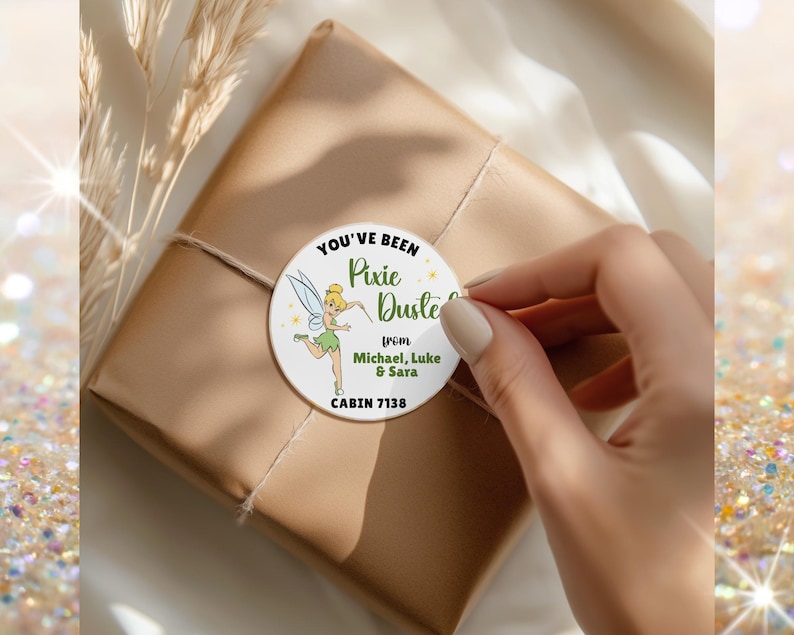 May include: A round white sticker featuring a cartoon fairy with blonde hair and green clothing, surrounded by yellow sparkles. The text reads: YOU'VE BEEN Pixie Dusted from Michael, Luke & Sara CABIN 7138. The sticker is applied to a brown paper package.
