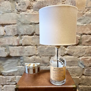 Könnte beinhalten: Eine Tischlampe aus einer durchsichtigen Glasflasche mit einem Etikett mit der Aufschrift "Monkey Shoulder The Original Blended Malt Scotch Whisky". Die Flasche hat einen schwarzen Metalldraht und eine dekorative Metallkette, die oben befestigt sind. Die Lampe hat einen weißen Stofflampenschirm.