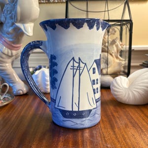 May include: A ceramic pitcher featuring a hand-painted blue and white nautical scene. The design depicts a sailboat on water next to a building, with a scalloped blue rim and a curved handle. The pitcher has a glossy finish and a cylindrical body.
