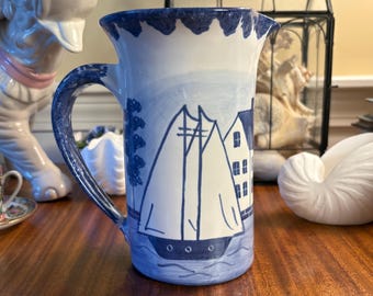 May include: A ceramic pitcher featuring a hand-painted blue and white nautical scene. The design depicts a sailboat on water next to a building, with a scalloped blue rim and a curved handle. The pitcher has a glossy finish and a cylindrical body.