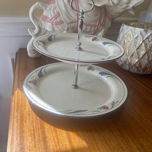 May include: A two-tiered serving stand featuring cream-colored ceramic plates with a floral pattern of blue, red, and green accents along the rims. The plates are connected by a central silver-toned metal rod with a looped handle at the top.