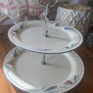 May include: Two-tiered serving tray with a silver handle. The white plates have a blue border and floral design in red, green, and blue. The tray is designed for serving desserts or appetizers.