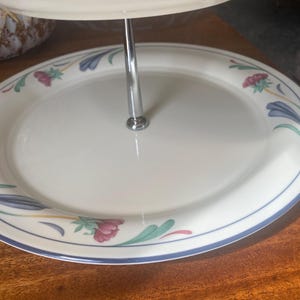 May include: A two-tiered serving platter with a white surface and a floral design. The rim is decorated with blue, green, and pink floral patterns. A silver metal rod connects the two tiers. The platter is on a wooden surface.