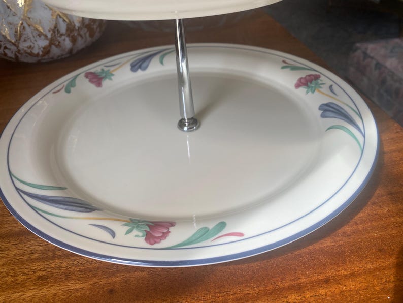 May include: A two-tiered serving platter with a white surface and a floral design. The rim is decorated with blue, green, and pink floral patterns. A silver metal rod connects the two tiers. The platter is on a wooden surface.