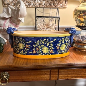 May include: An oval ceramic planter featuring a deep blue background with a repeating pattern of yellow fruit and green vines. The rim and base are painted in a solid yellow hue. Two dark blue handles shaped like ram heads are on the sides.